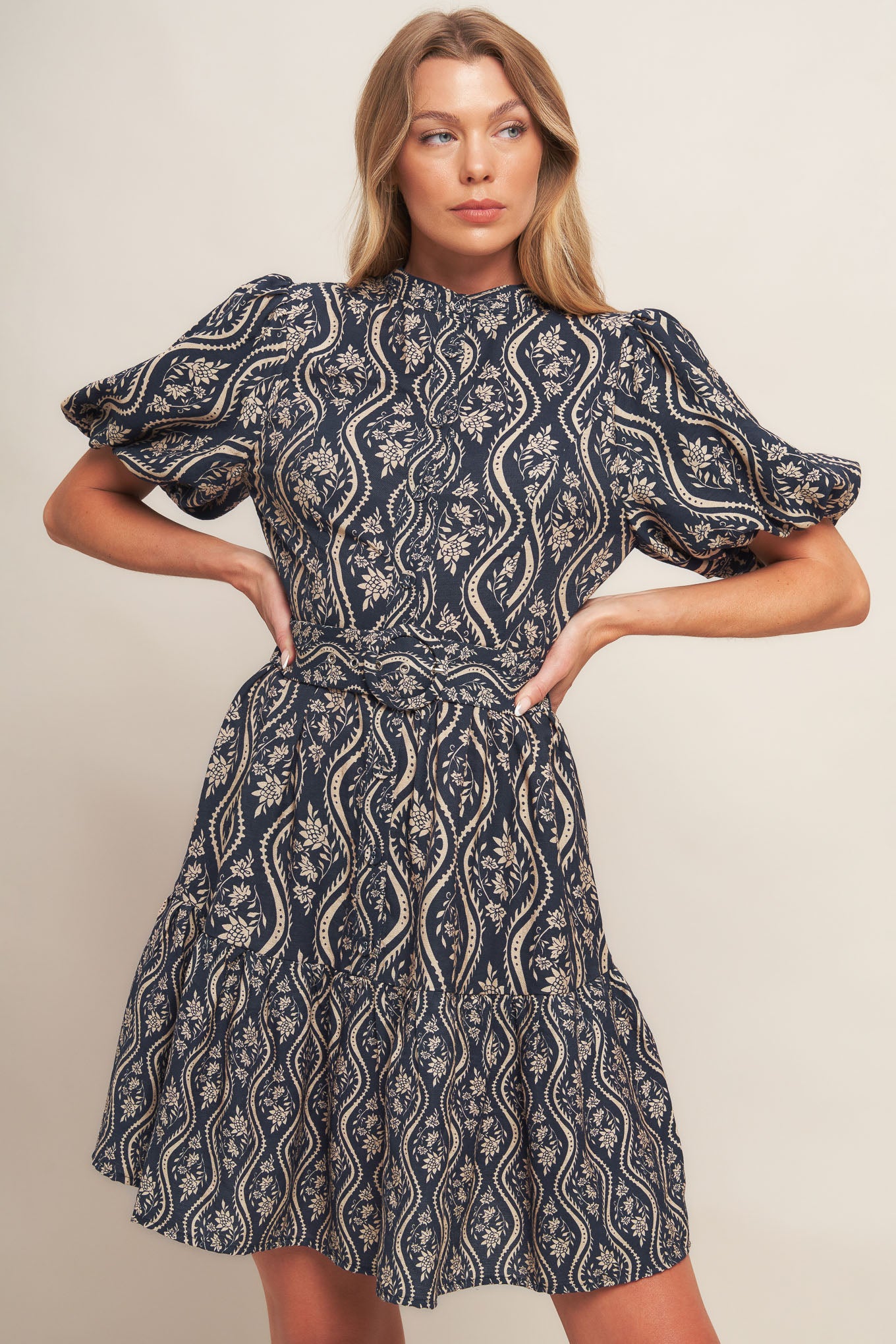 A printed woven mini dress featuring high neckline, short puff sleeve, front button down closure, self belt and ruffled hemline