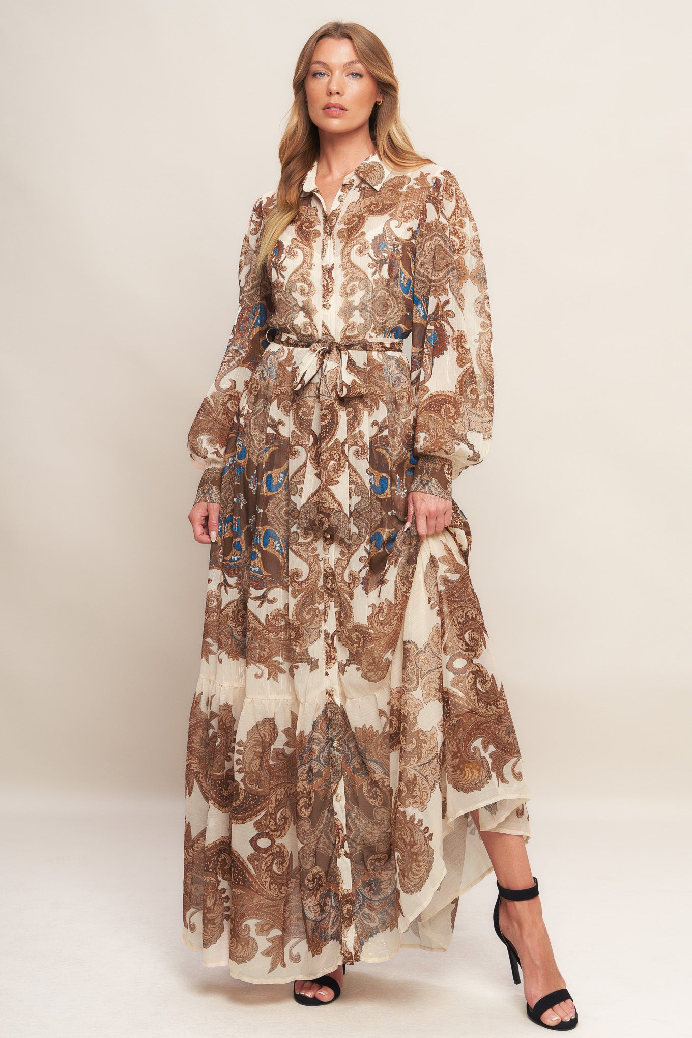 A printed woven metallic threaded maxi dress featuring collar, long sleeve with cuff, self sash tie, front button down closure and ruffled hemline