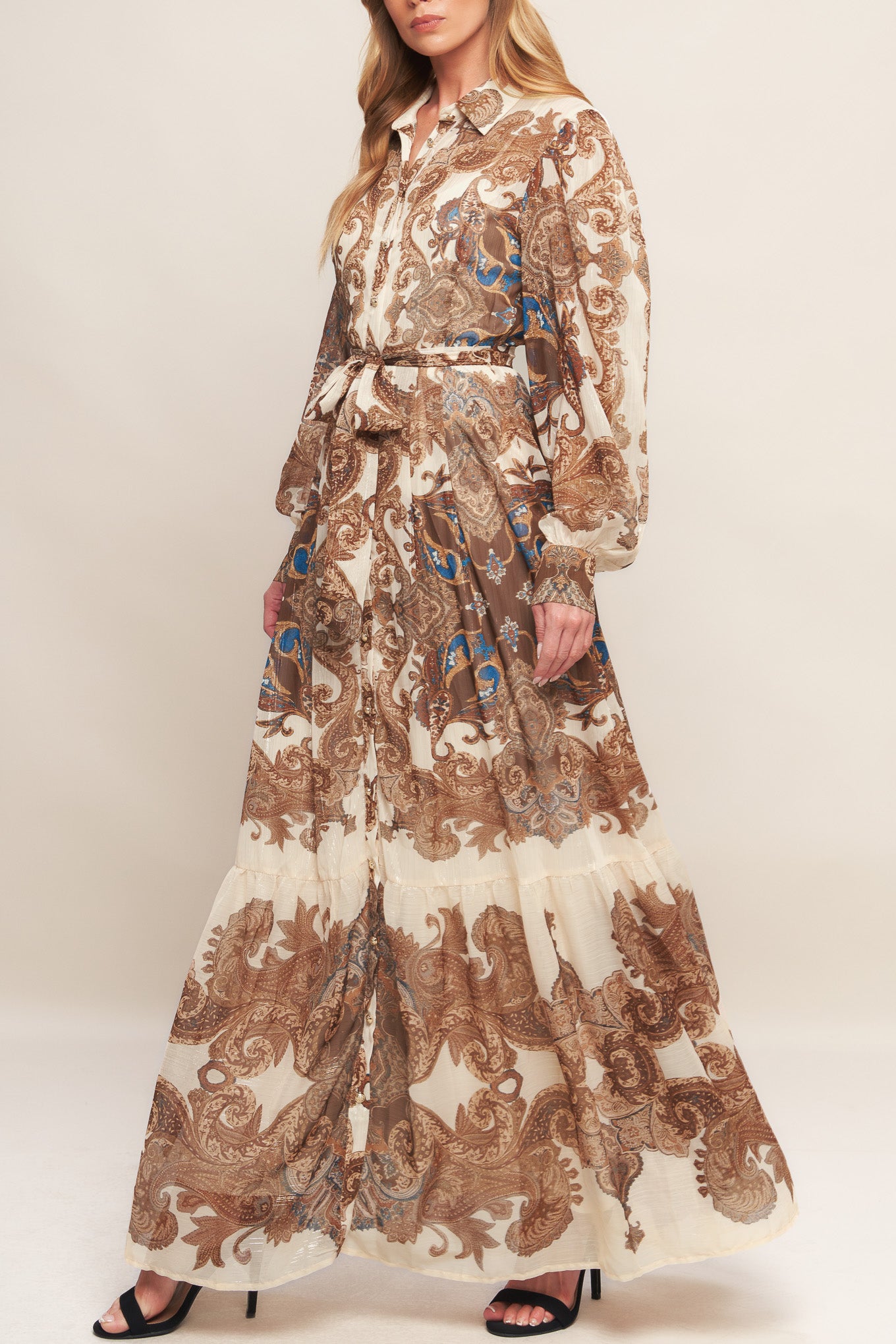 A printed woven metallic threaded maxi dress featuring collar, long sleeve with cuff, self sash tie, front button down closure and ruffled hemline