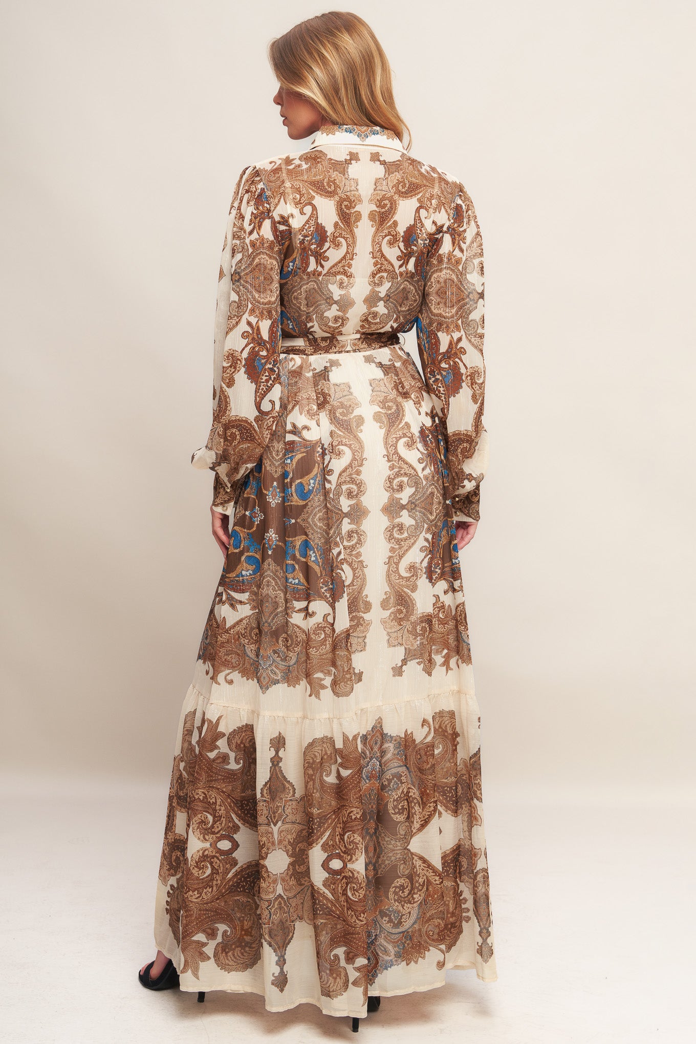 A printed woven metallic threaded maxi dress featuring collar, long sleeve with cuff, self sash tie, front button down closure and ruffled hemline