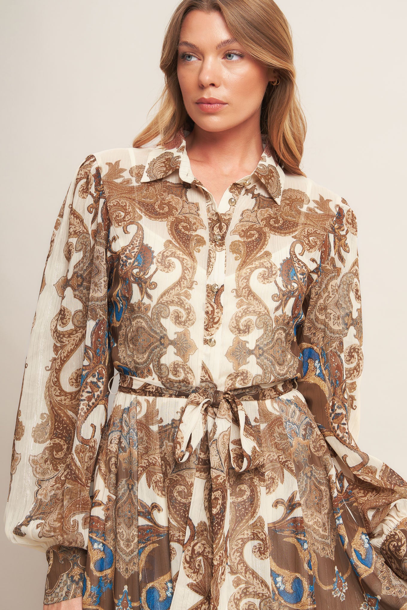 A printed woven metallic threaded maxi dress featuring collar, long sleeve with cuff, self sash tie, front button down closure and ruffled hemline