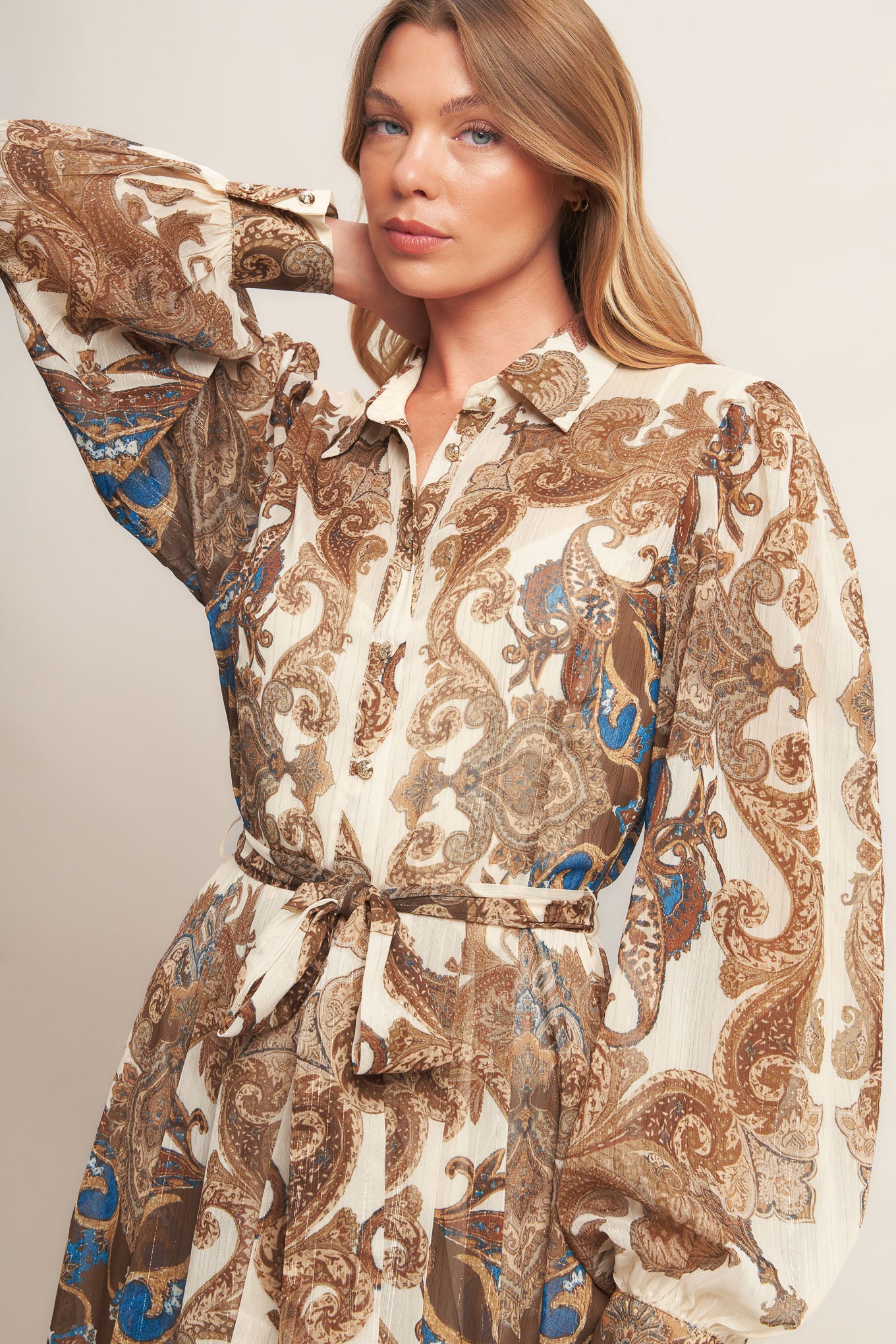 A printed woven metallic threaded maxi dress featuring collar, long sleeve with cuff, self sash tie, front button down closure and ruffled hemline