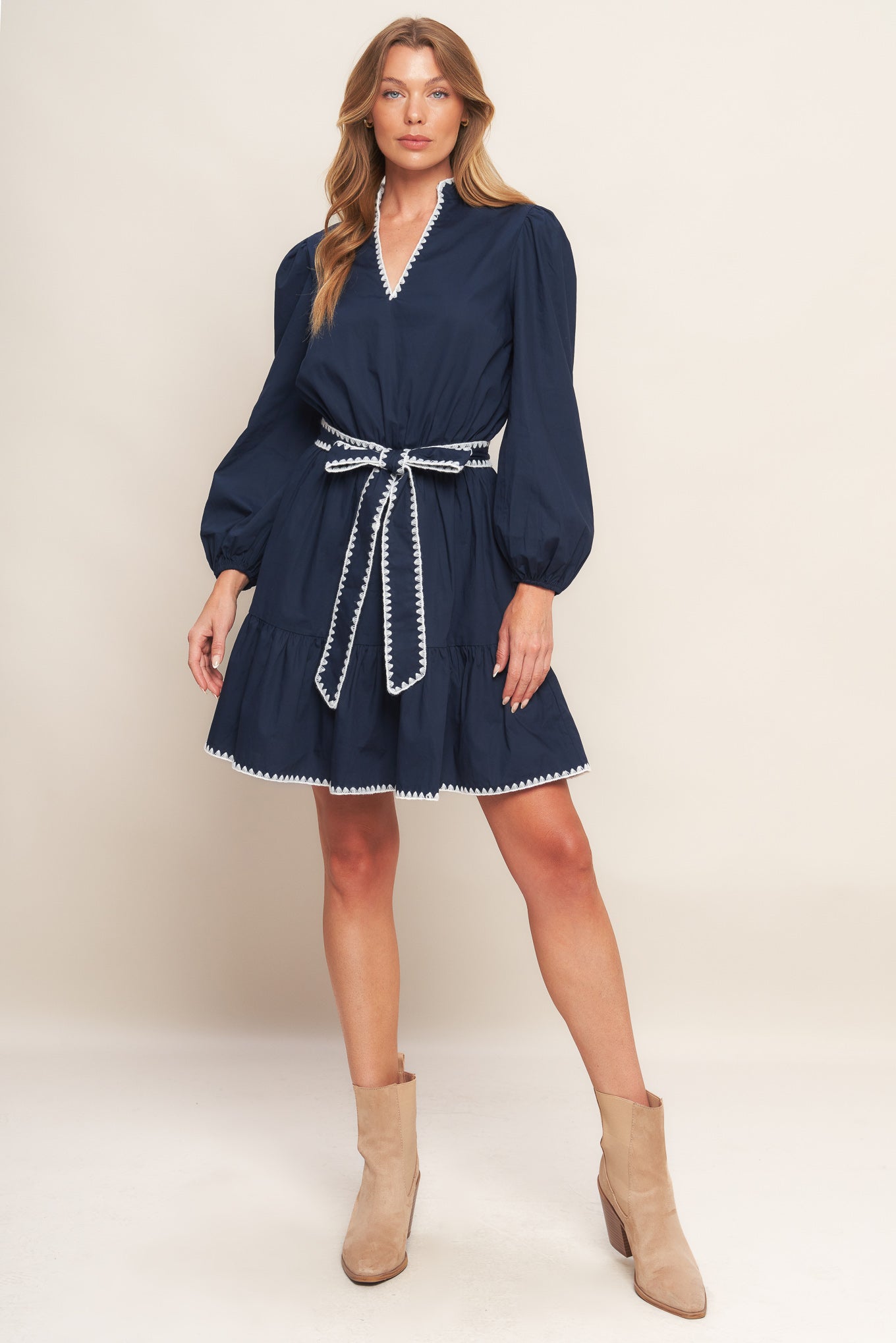 A solid woven mini dress featuring V neckline, long sleeve, elasticized waist, self sash tie, ruffled hemline and embroidered edge detail