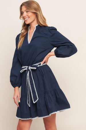 A solid woven mini dress featuring V neckline, long sleeve, elasticized waist, self sash tie, ruffled hemline and embroidered edge detail