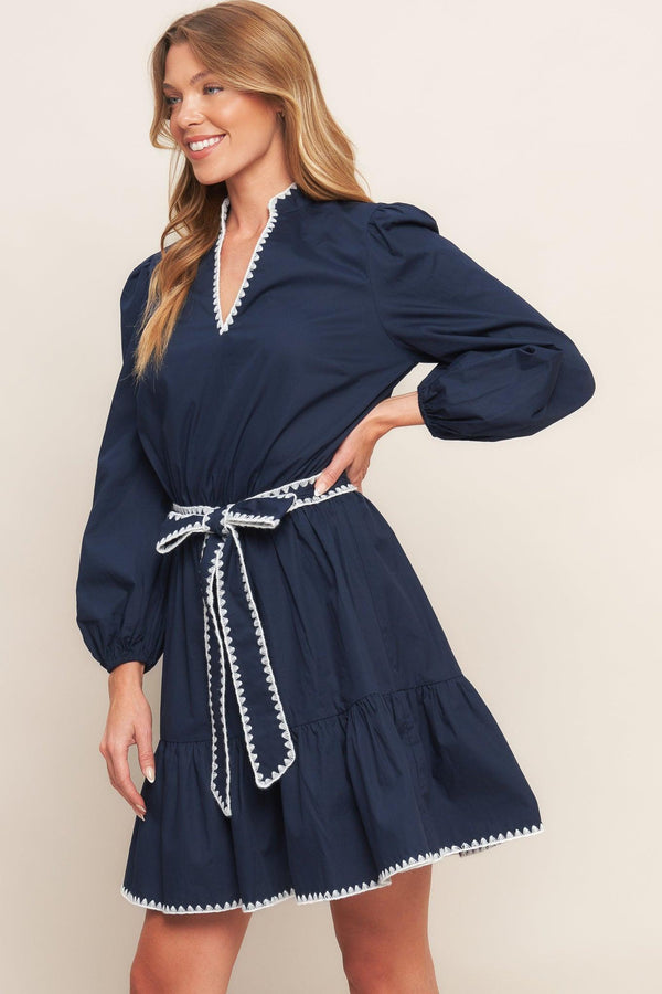 A solid woven mini dress featuring V neckline, long sleeve, elasticized waist, self sash tie, ruffled hemline and embroidered edge detail