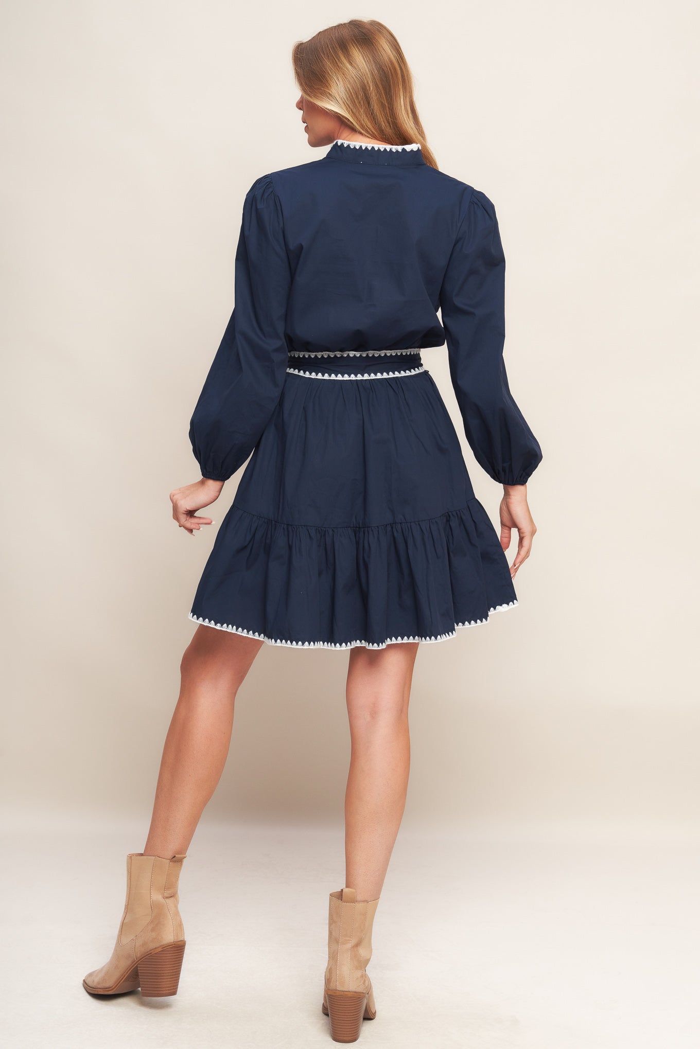 A solid woven mini dress featuring V neckline, long sleeve, elasticized waist, self sash tie, ruffled hemline and embroidered edge detail