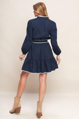 A solid woven mini dress featuring V neckline, long sleeve, elasticized waist, self sash tie, ruffled hemline and embroidered edge detail