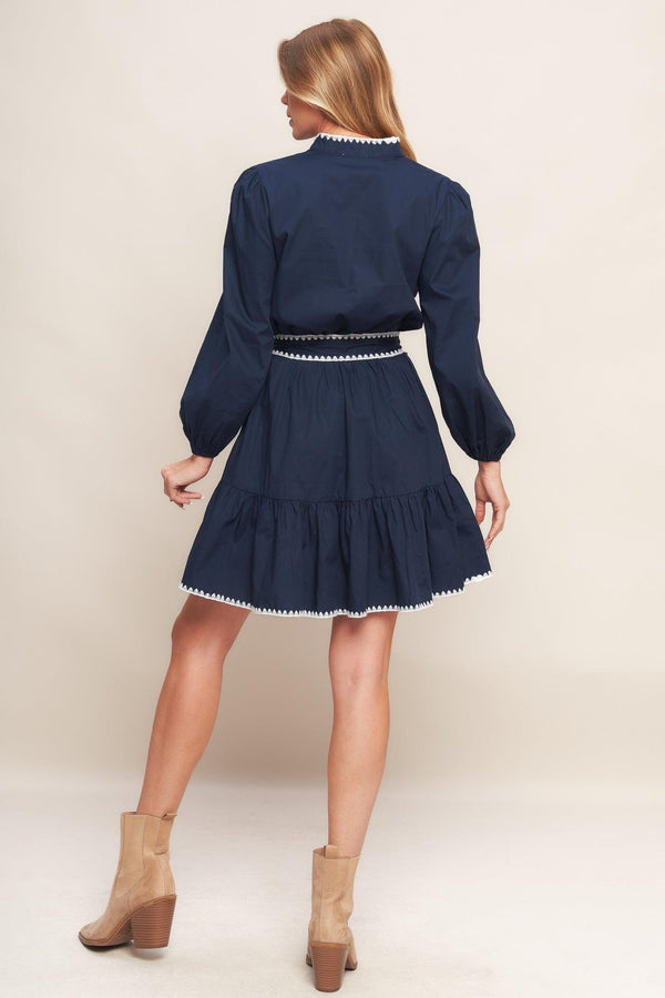 A solid woven mini dress featuring V neckline, long sleeve, elasticized waist, self sash tie, ruffled hemline and embroidered edge detail