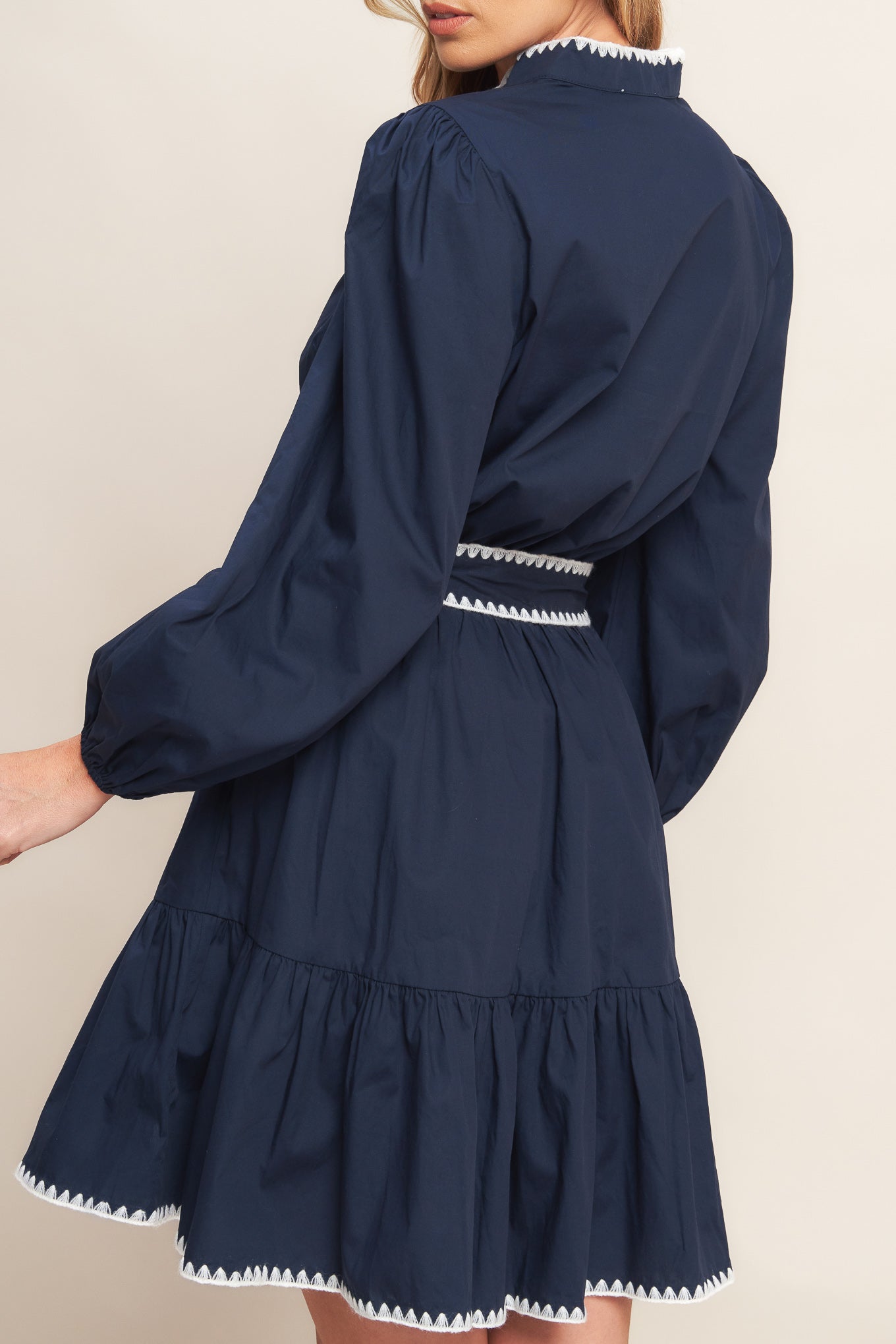 A solid woven mini dress featuring V neckline, long sleeve, elasticized waist, self sash tie, ruffled hemline and embroidered edge detail