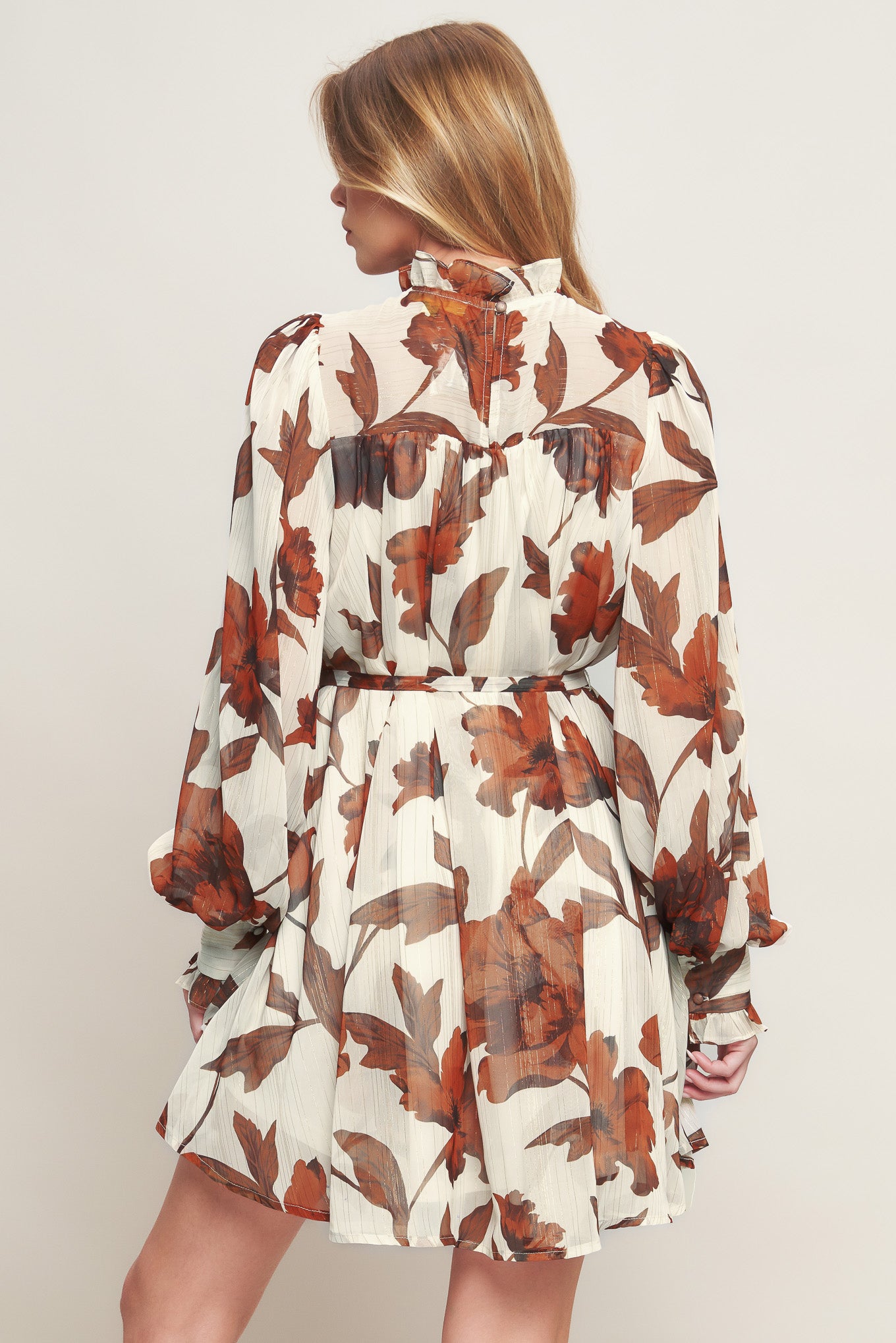 A printed woven mini dress featuring high ruffled neckline, long sleeve with cuff, self sash tie and back neck button closure