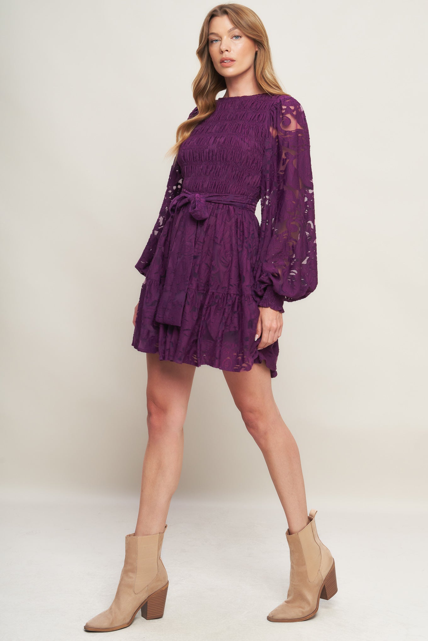 A woven lace mini dress featuring round neckline, long sleeve with cuff, smocked bodice, self sash tie, ruffled hemline and back neck button closure