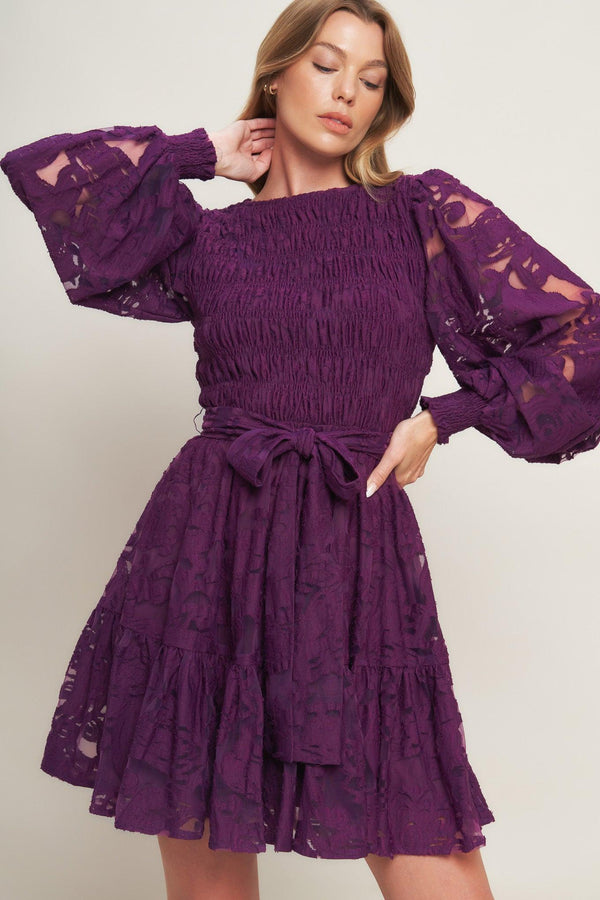 A woven lace mini dress featuring round neckline, long sleeve with cuff, smocked bodice, self sash tie, ruffled hemline and back neck button closure