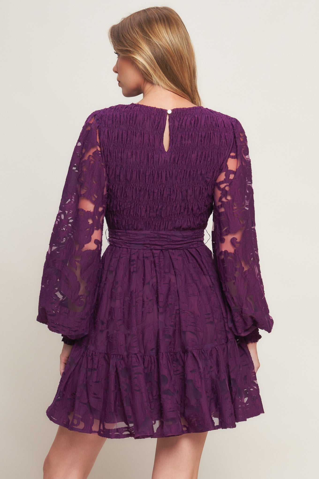 A woven lace mini dress featuring round neckline, long sleeve with cuff, smocked bodice, self sash tie, ruffled hemline and back neck button closure