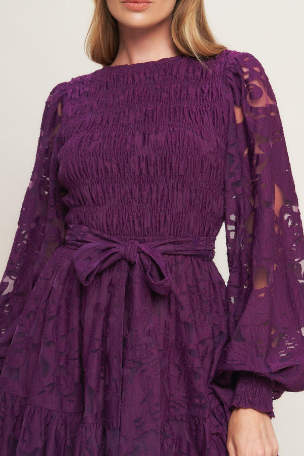 A woven lace mini dress featuring round neckline, long sleeve with cuff, smocked bodice, self sash tie, ruffled hemline and back neck button closure