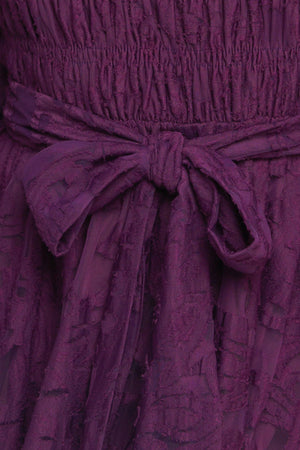 A woven lace mini dress featuring round neckline, long sleeve with cuff, smocked bodice, self sash tie, ruffled hemline and back neck button closure