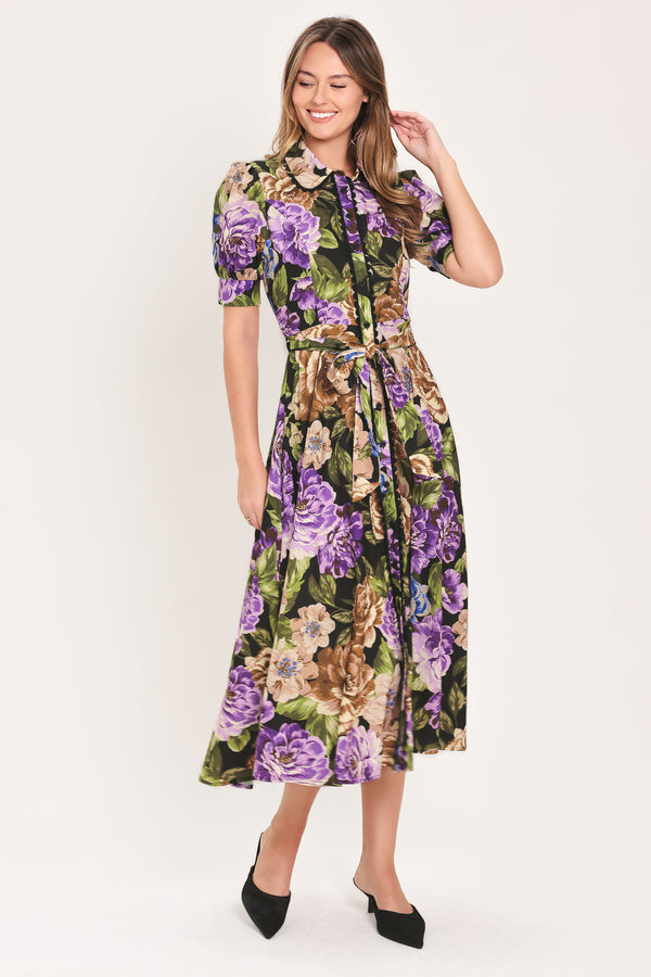 Woman wearing a black short sleeve, collared, button shirt dress with purple and gold floral pattern and a self tie waist belt | Flying Tomato