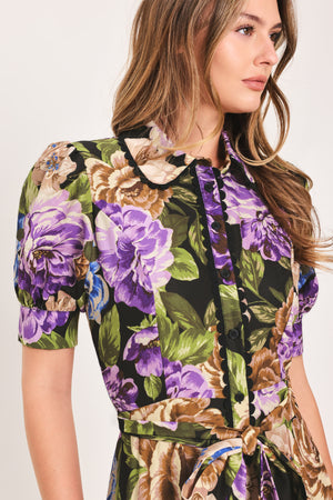 Woman wearing a black short sleeve, collared, button shirt dress with purple and gold floral pattern and a self tie waist belt | Flying Tomato