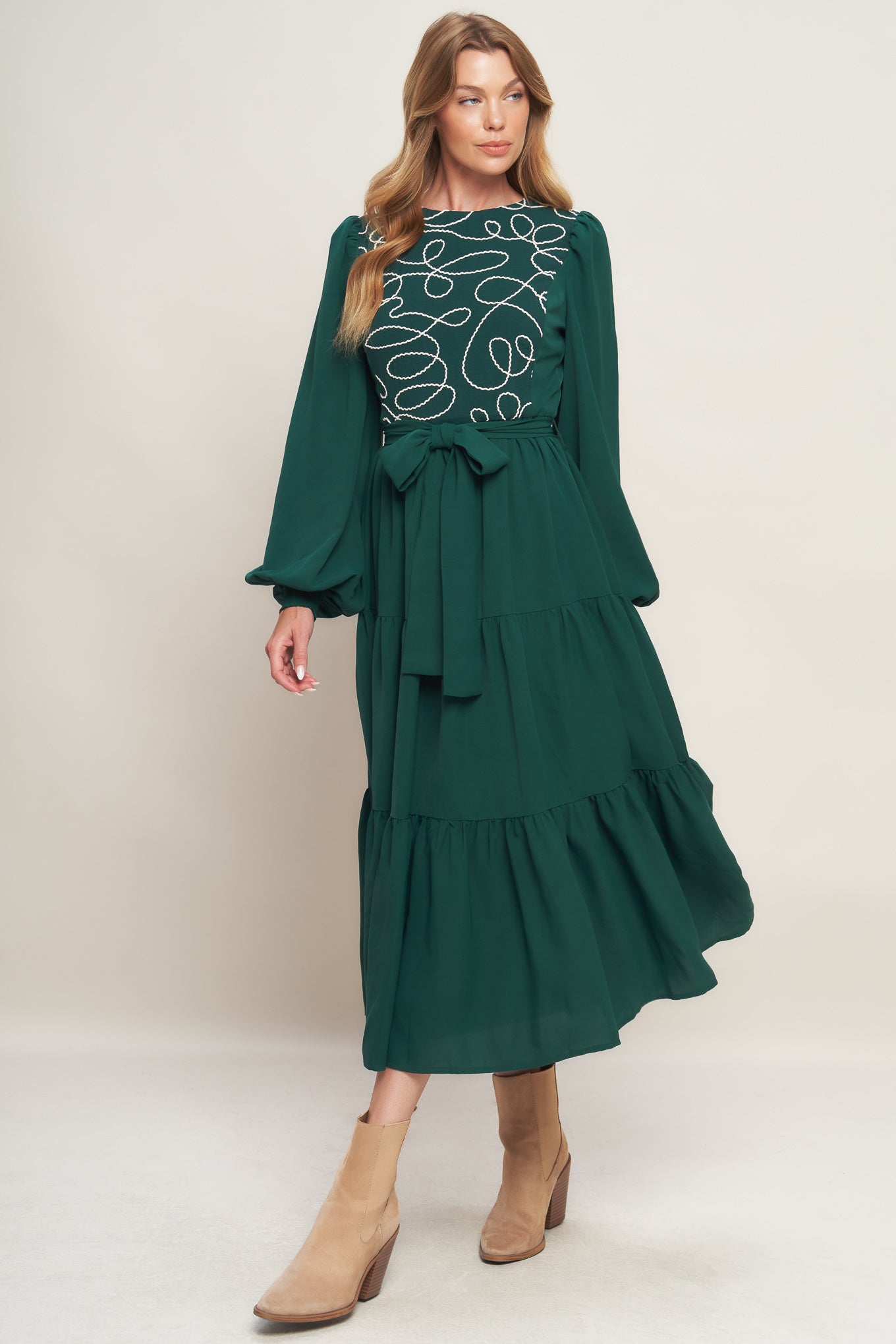 A solid woven midi dress featuring round neckline, long sleeve with smocked cuff, tiered skirt with self sash tie and back zipper closure