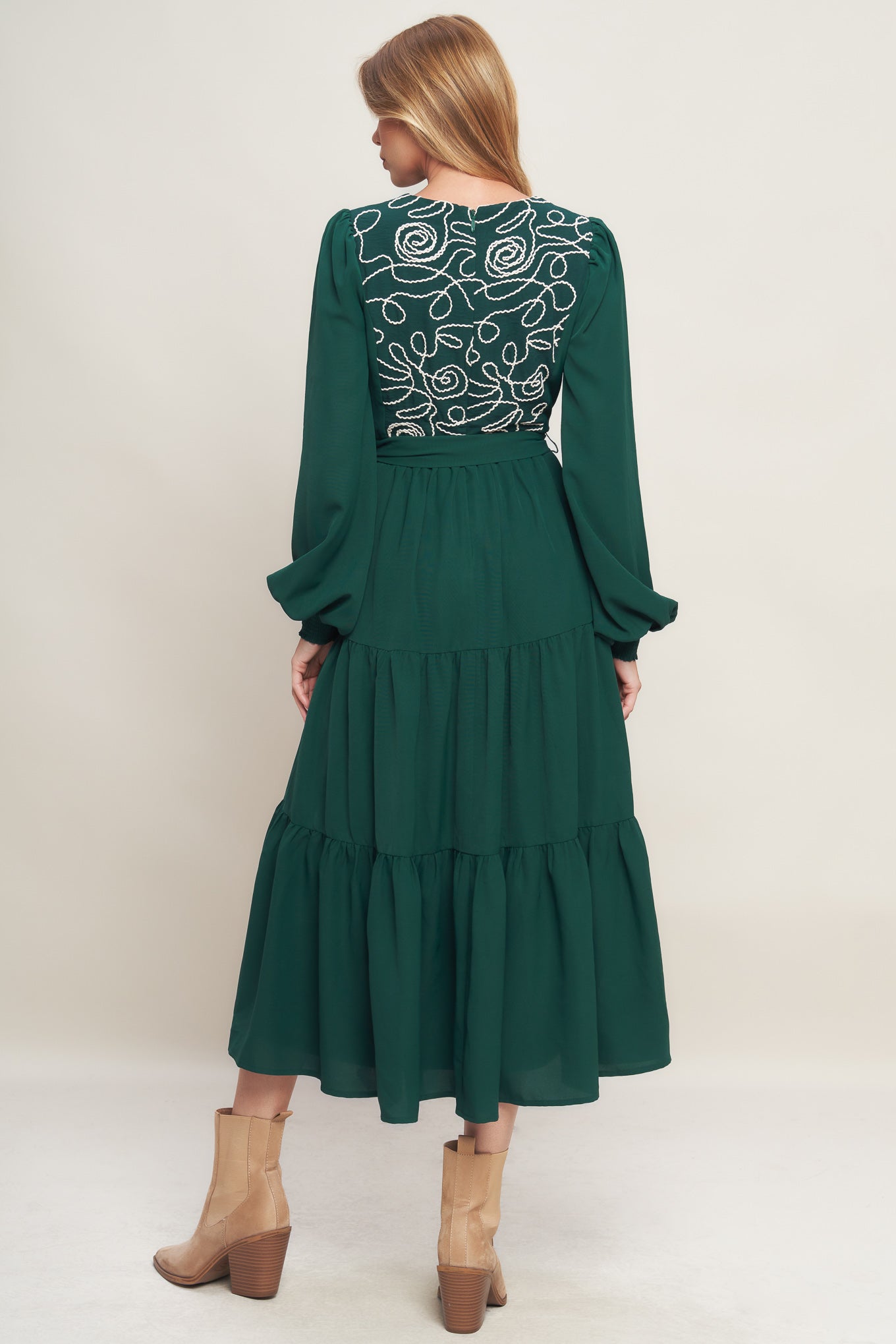 A solid woven midi dress featuring round neckline, long sleeve with smocked cuff, tiered skirt with self sash tie and back zipper closure