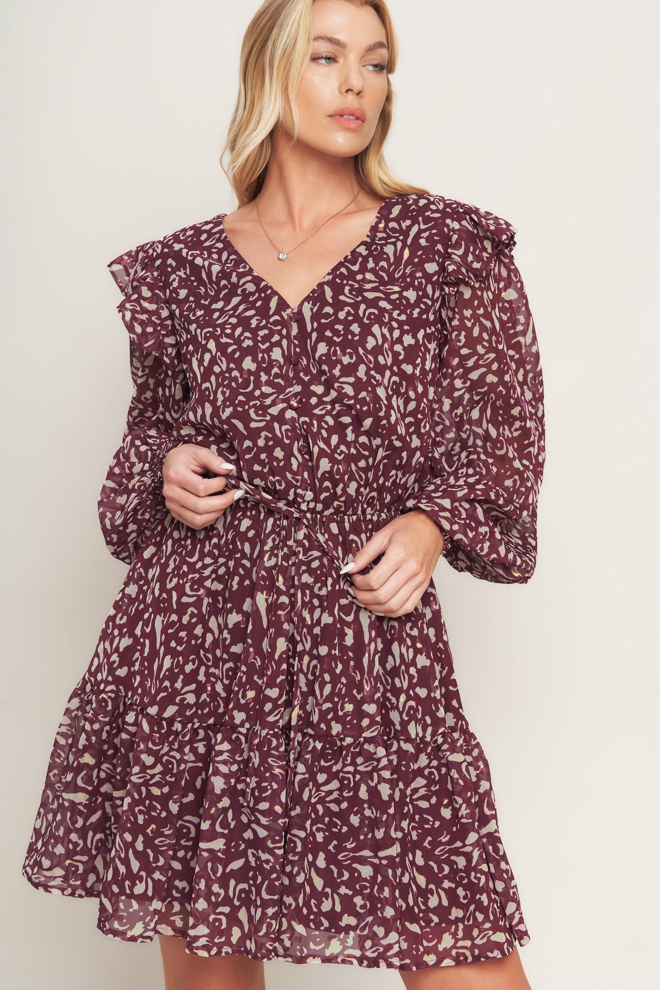 A printed woven mini dress featuring V neckline, long sleeve with ruffled shoulder, front button detail, elasticized waist with front tie and ruffled hemline