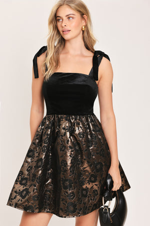 Woman wearing a sleeveless mini dress with a black velvet square neck top with lace up shoulder straps and bronze circle skirt with black jacquard flowers | Flying Tomato