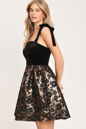 Woman wearing a sleeveless mini dress with a black velvet square neck top with lace up shoulder straps and bronze circle skirt with black jacquard flowers | Flying Tomato