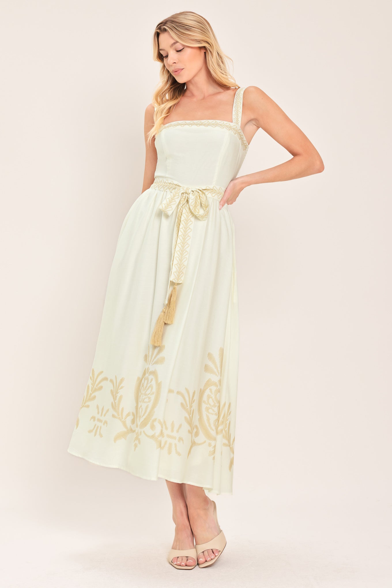 Woman wearing a ivory sleeveless dress with gold embroidery featuring straight neckline, straps, self sash tie with tassel, full skirt and smocked back bodice | Flying Tomato