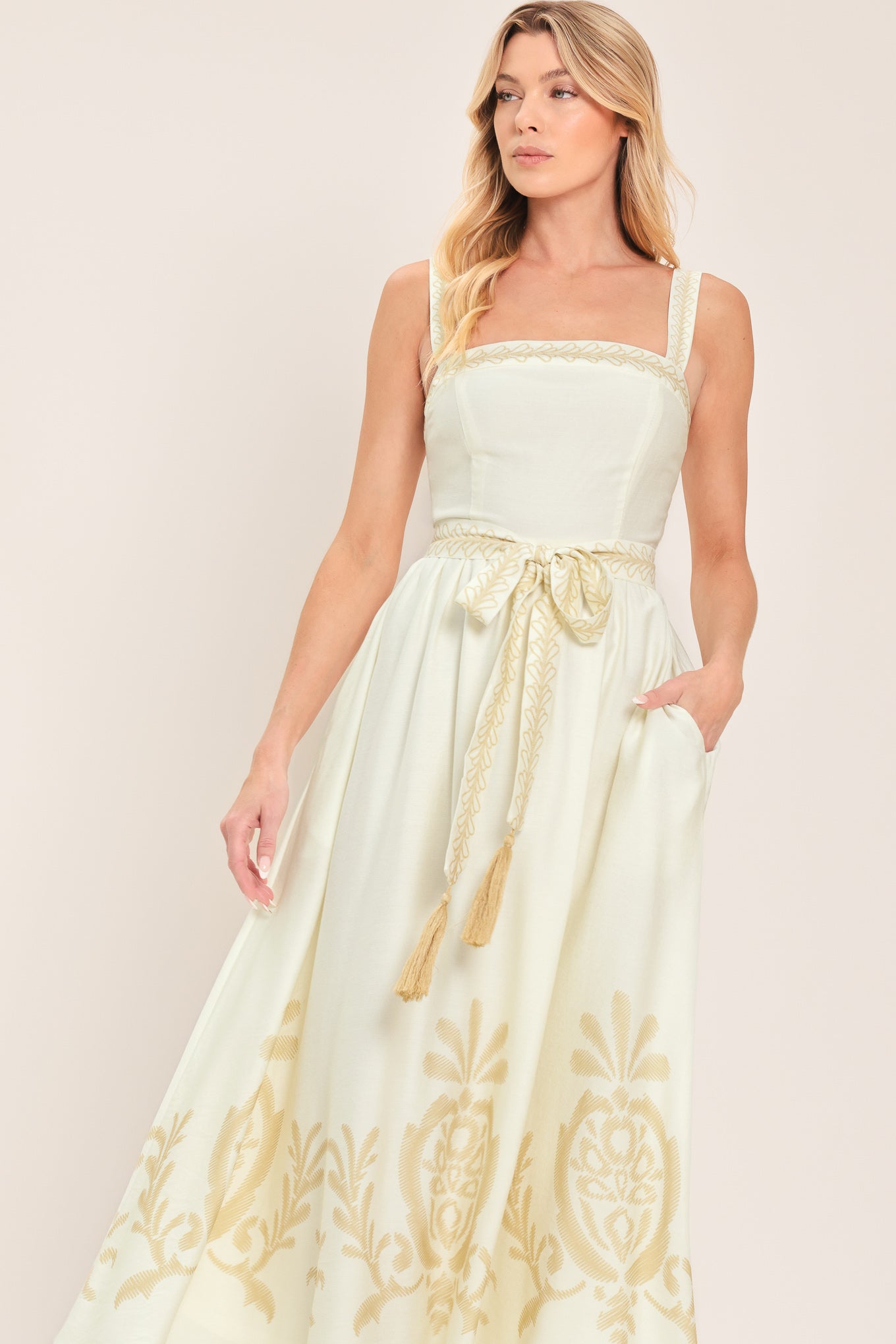 Woman wearing a ivory sleeveless dress with gold embroidery featuring straight neckline, straps, self sash tie with tassel, full skirt and smocked back bodice | Flying Tomato