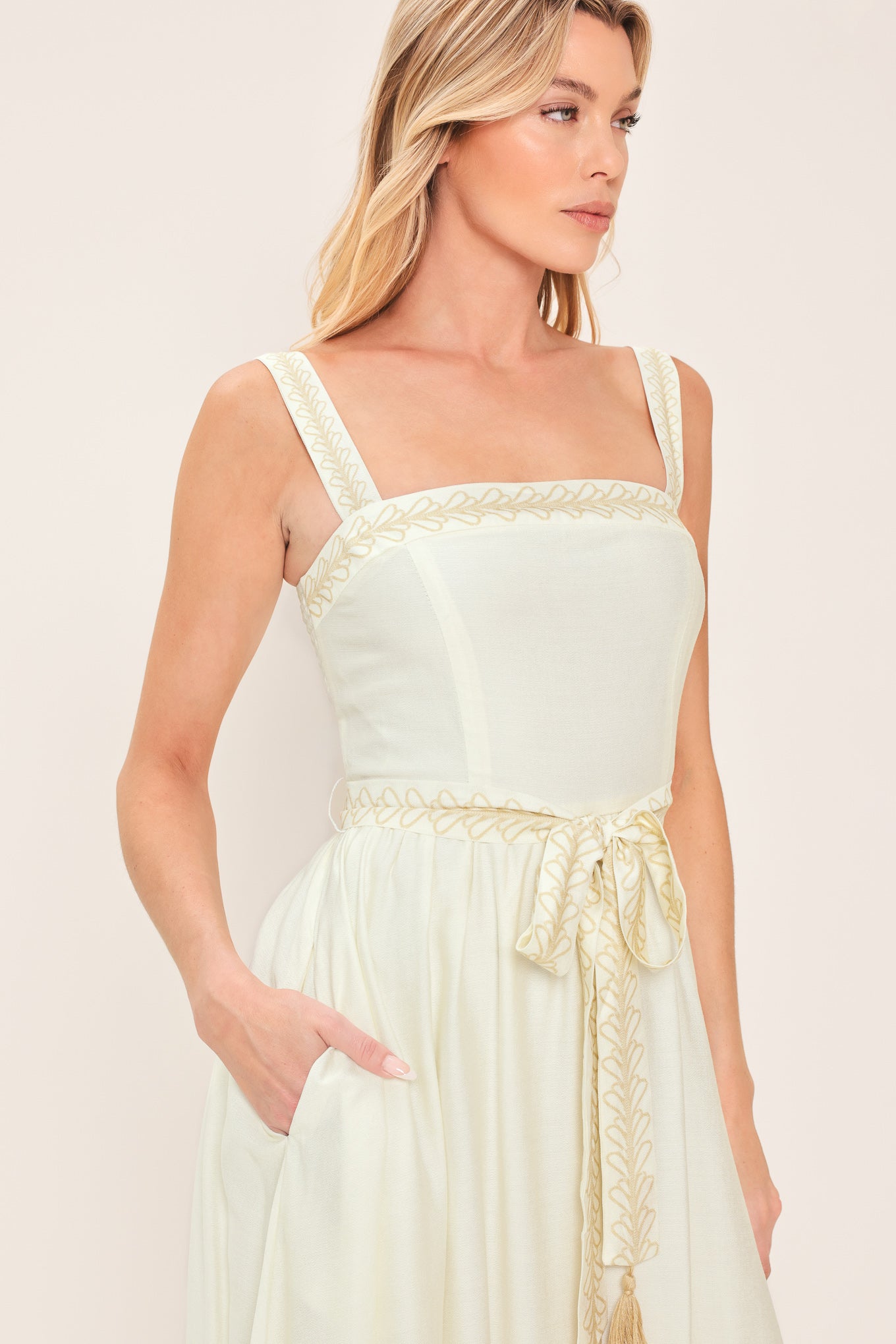 Woman wearing a ivory sleeveless dress with gold embroidery featuring straight neckline, straps, self sash tie with tassel, full skirt and smocked back bodice | Flying Tomato