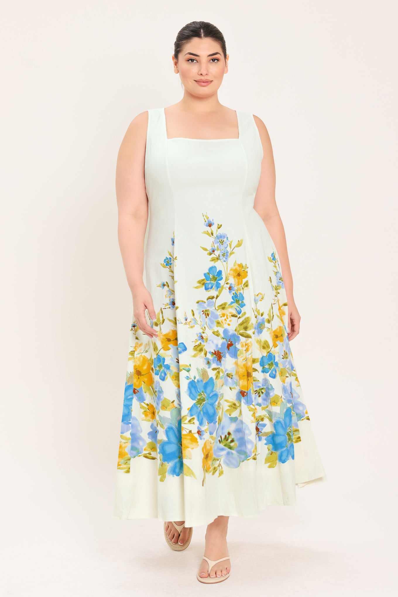 A woman modeling a sleeveless midi dress with a square neckline and a white base featuring a blue and yellow floral print on the skirt | Plus Curve Flying Tomato