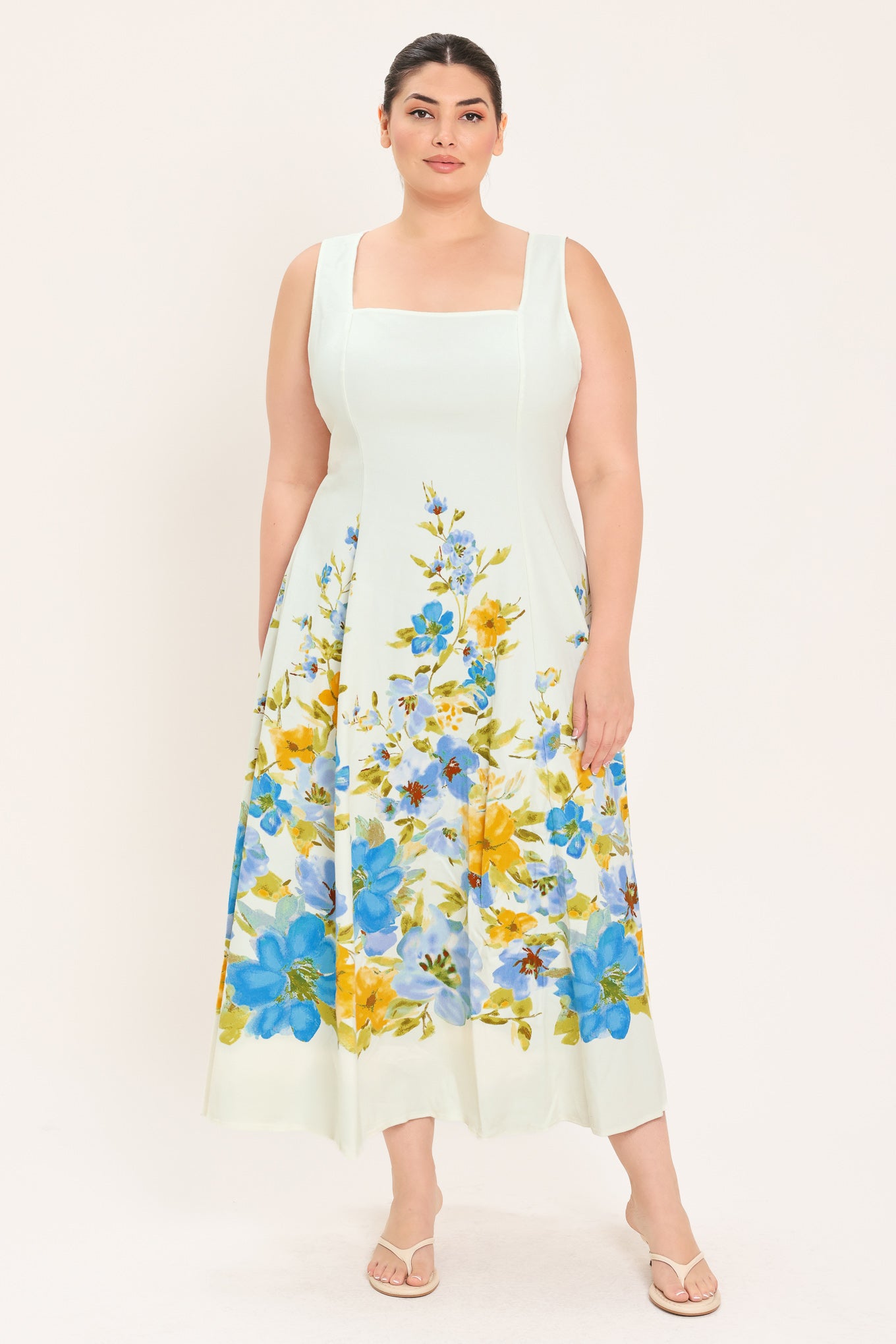 A woman modeling a sleeveless midi dress with a square neckline and a white base featuring a blue and yellow floral print on the skirt | Plus Curve Flying Tomato