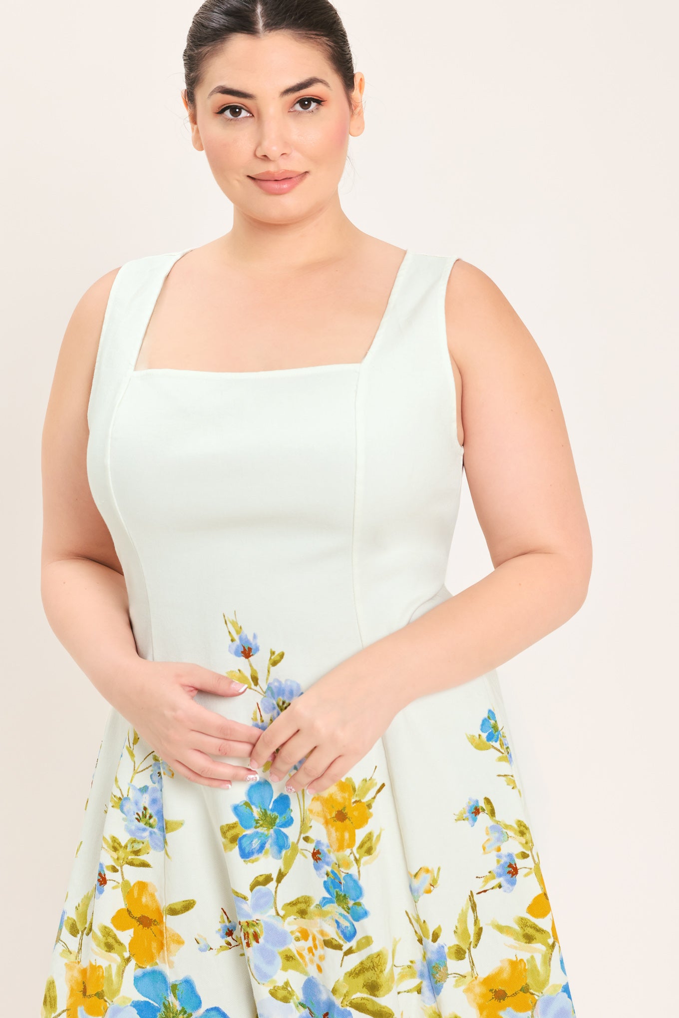 A woman modeling a sleeveless midi dress with a square neckline and a white base featuring a blue and yellow floral print on the skirt | Plus Curve Flying Tomato