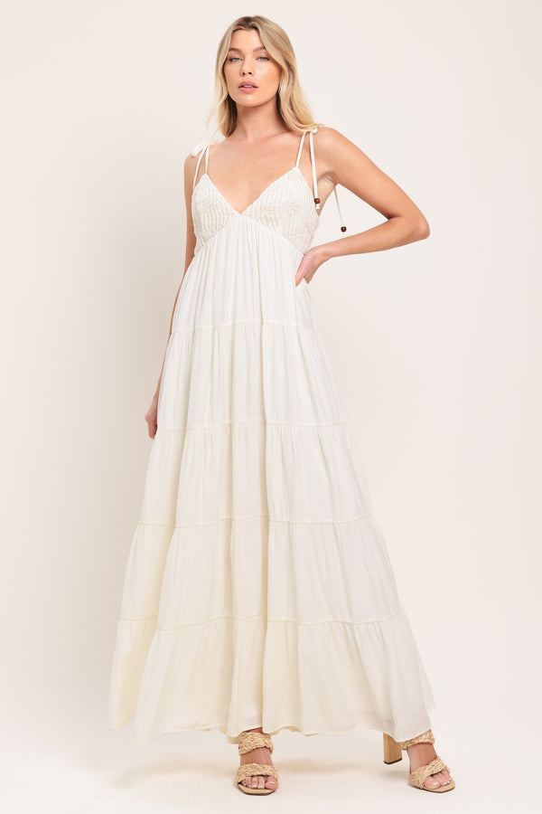 Woman wearing a cream flowy maxi dress featuring lace bodice, shoulder tie, tier body and smocked back bodice | Flying Tomato
