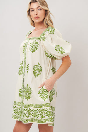 Woman wearing a white printed woven mini dress with green embroidery featuring a square neckline, pockets, short puff sleeve and front button detail on a white background | Flying Tomato