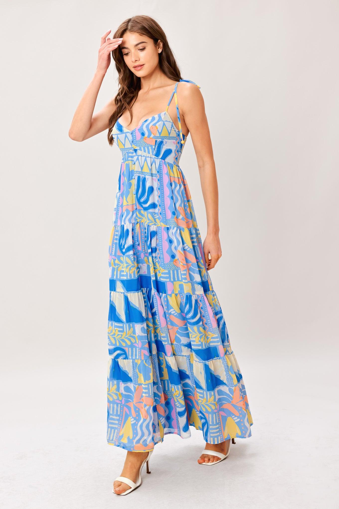 Woman wearing a colorful geometric patterned printed woven maxi dress featuring cami top with shoulder tie, front button detail, tier skirt and smocked back bodice