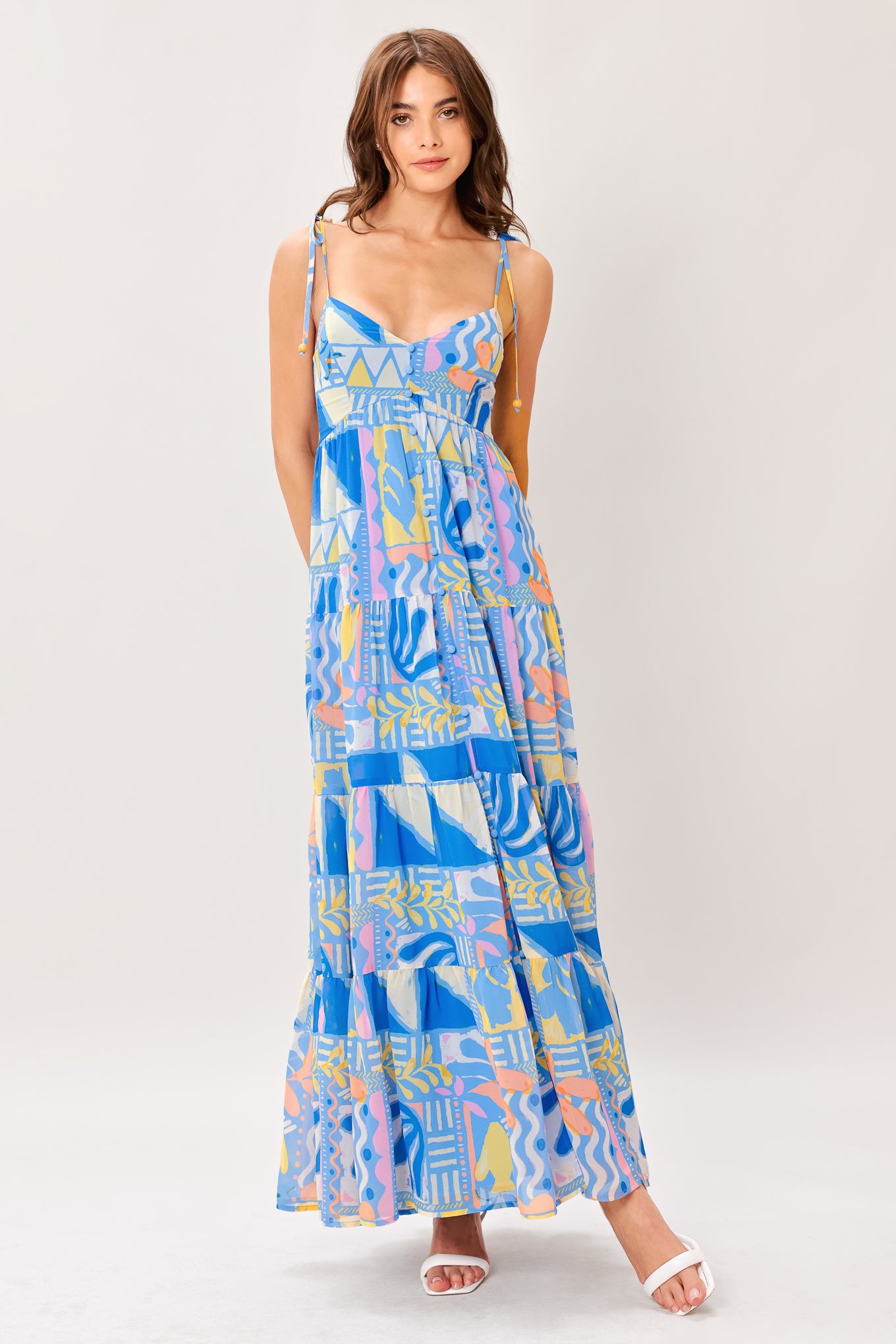 Woman wearing a colorful geometric patterned printed woven maxi dress featuring cami top with shoulder tie, front button detail, tier skirt and smocked back bodice