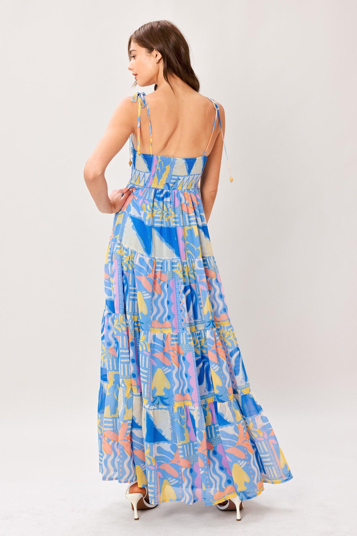 Woman wearing a colorful geometric patterned printed woven maxi dress featuring cami top with shoulder tie, front button detail, tier skirt and smocked back bodice