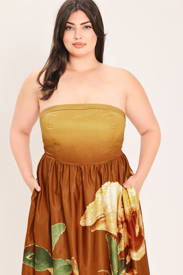 A woman modeling a strapless dress with a mustard yellow bandeau-style bodice and a brown skirt featuring a large green and gold floral print, set against a plain light background.