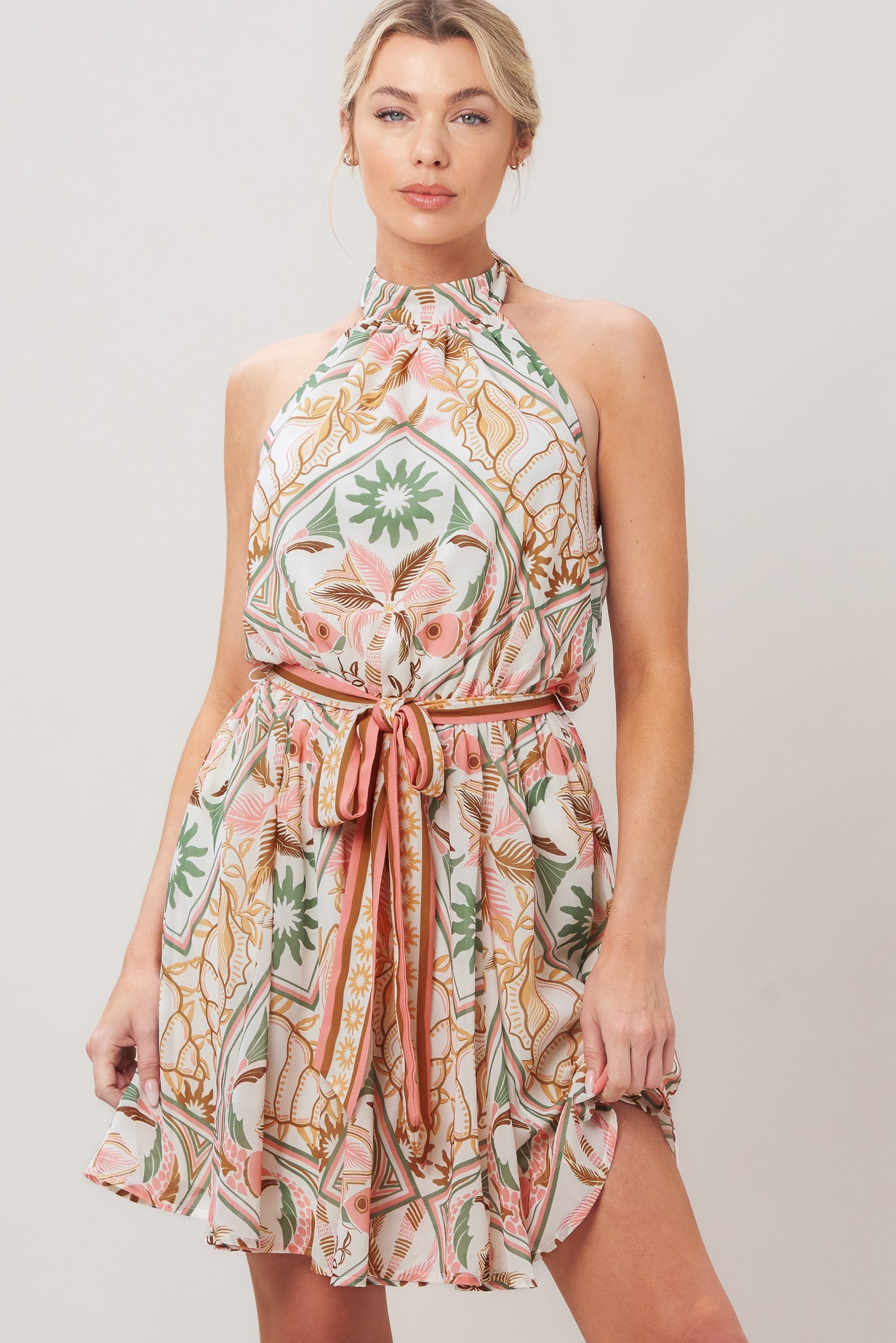 Woman wearing a printed woven mini dress featuring halter neckline, elastic waist with self sash, full skirt, bare back with tie on a white background | Flying Tomato