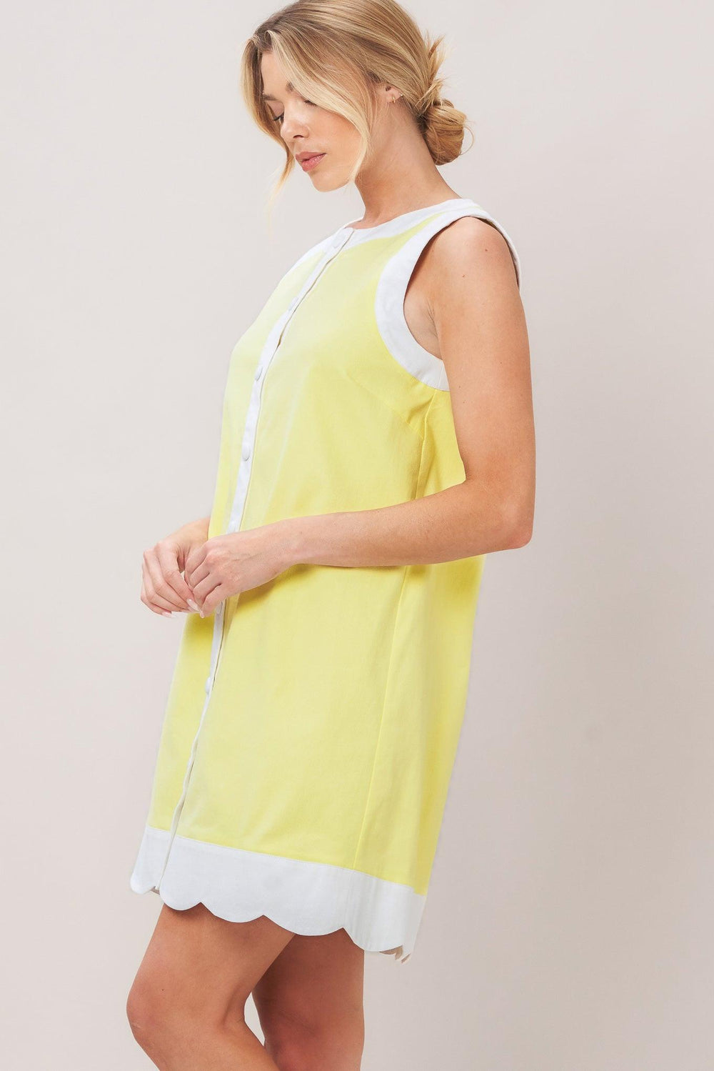 Woman wearing a pink with white trim mini dress featuring round neckline, sleeveless, front button down and scallop hem