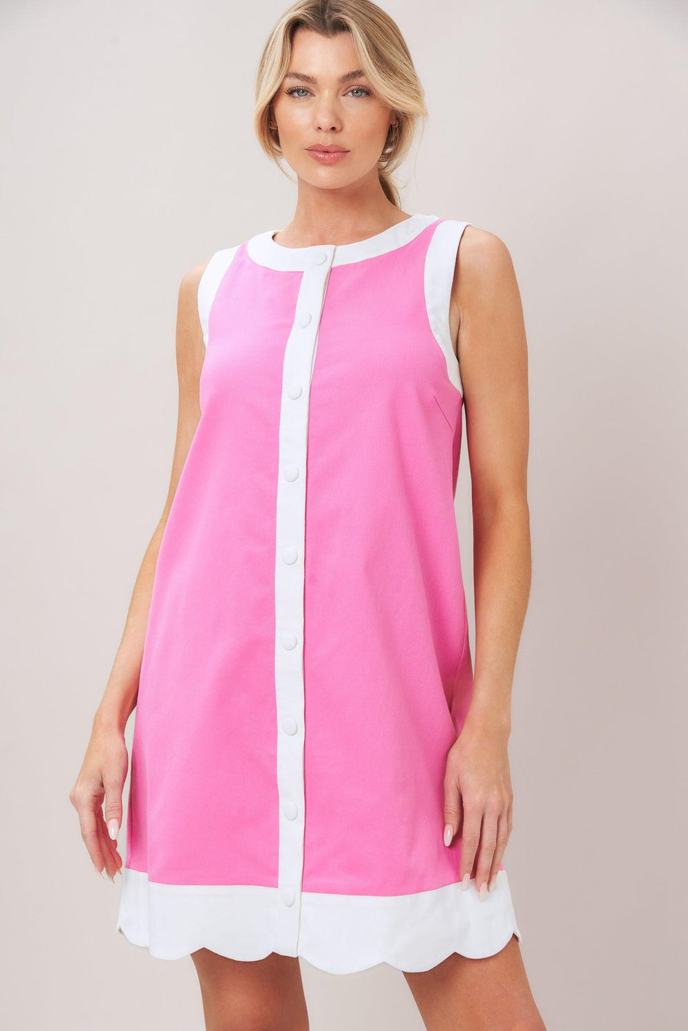 Woman wearing a pink with white trim mini dress featuring round neckline, sleeveless, front button down and scallop hem