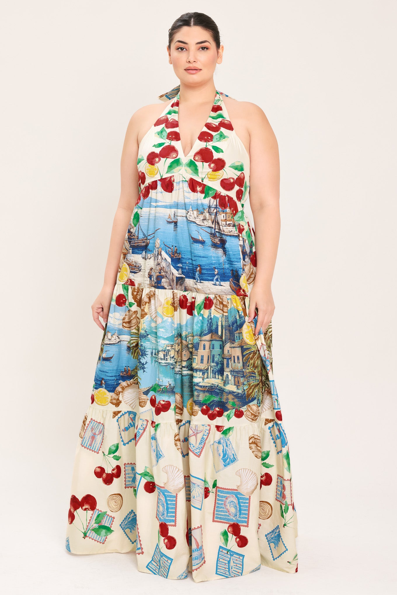 A woman modeling a halter-neck maxi dress with a colorful cherry and coastal harbor print on a cream background | Plus Curve Flying Tomato