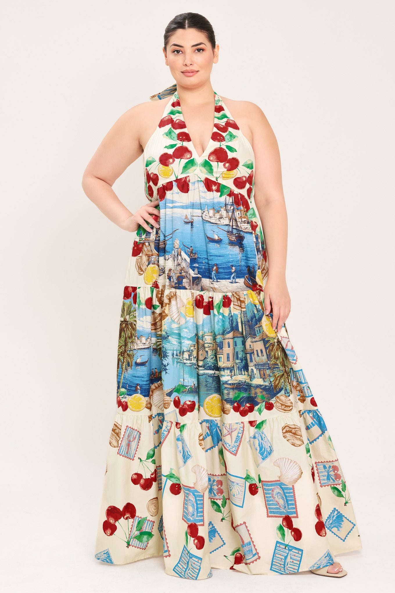 A woman modeling a halter-neck maxi dress with a colorful cherry and coastal harbor print on a cream background | Plus Curve Flying Tomato