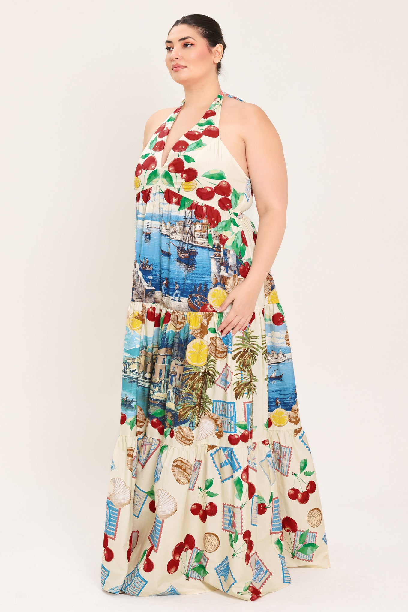 A woman modeling a halter-neck maxi dress with a colorful cherry and coastal harbor print on a cream background | Plus Curve Flying Tomato