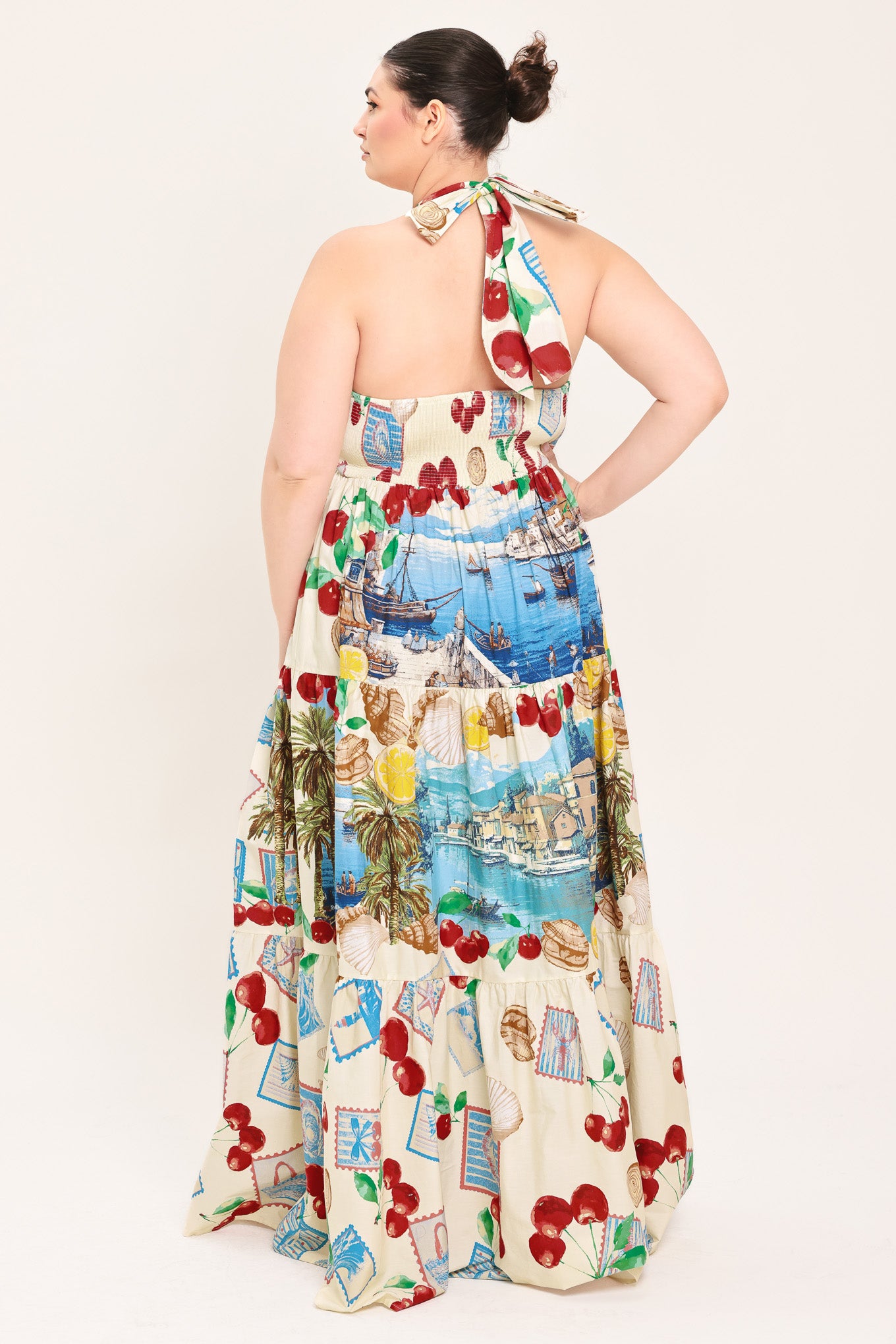 A woman modeling a halter-neck maxi dress with a colorful cherry and coastal harbor print on a cream background | Plus Curve Flying Tomato