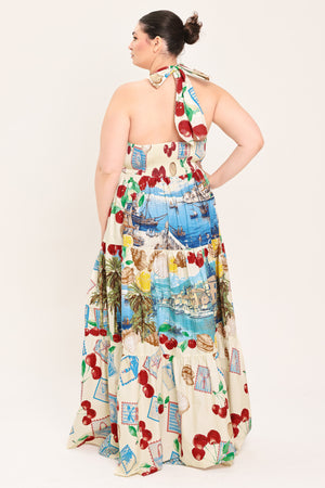 A woman modeling a halter-neck maxi dress with a colorful cherry and coastal harbor print on a cream background | Plus Curve Flying Tomato