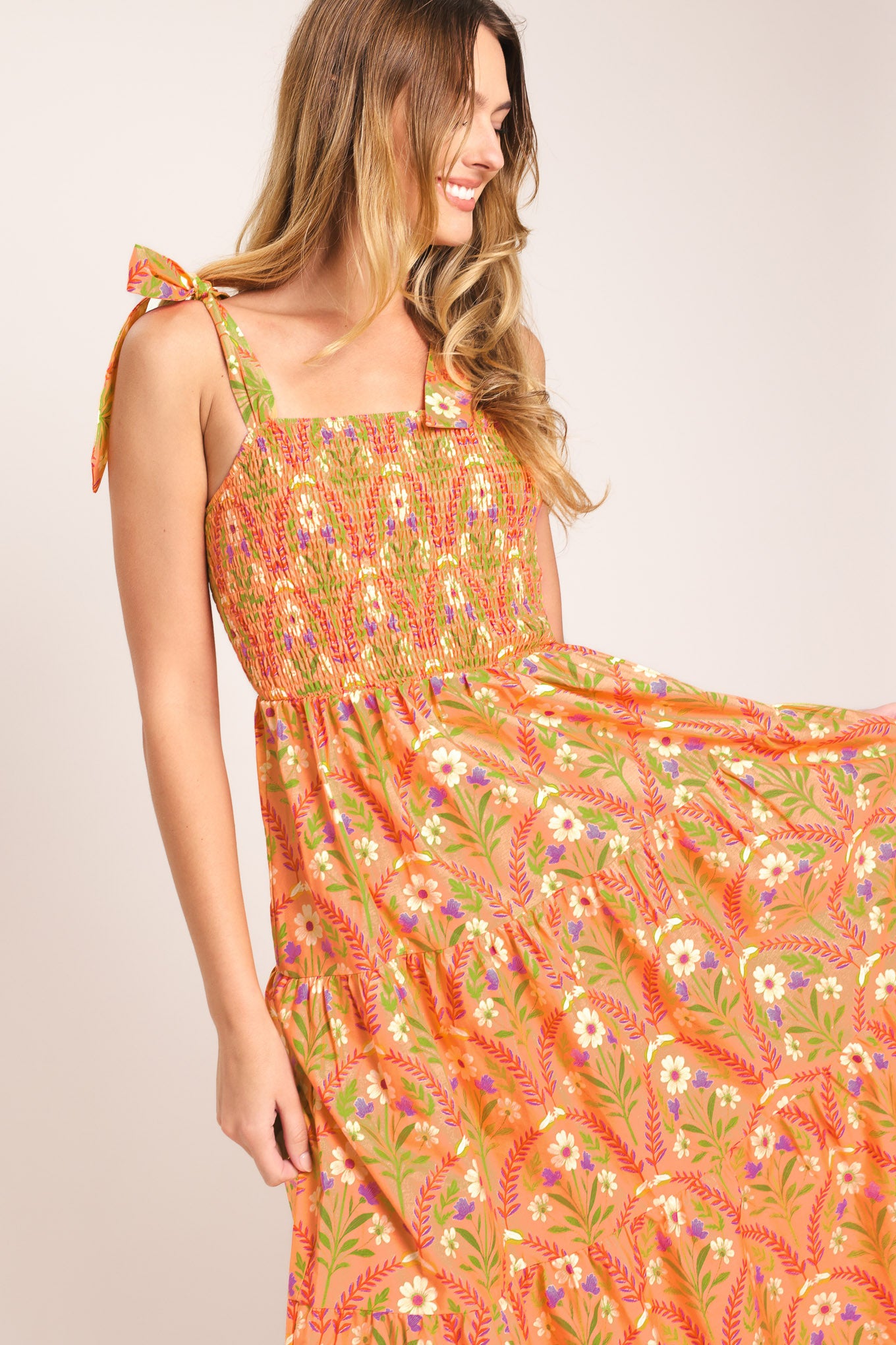 Woman wearing a floral patterned orange maxi dress featuring straight neckline, smocked bodice, shoulder ties and tier skirt | Flying Tomato