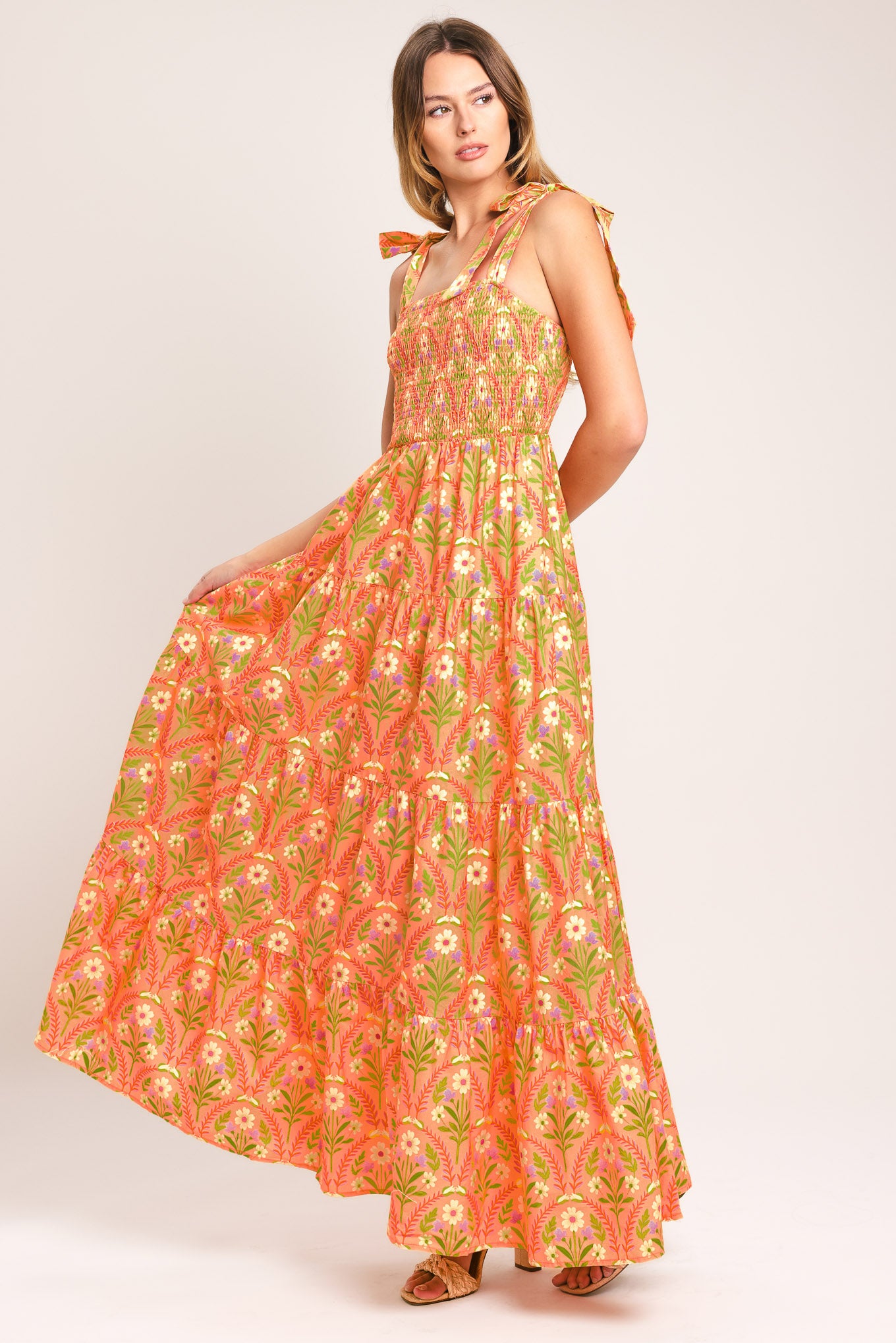 Woman wearing a floral patterned orange maxi dress featuring straight neckline, smocked bodice, shoulder ties and tier skirt | Flying Tomato