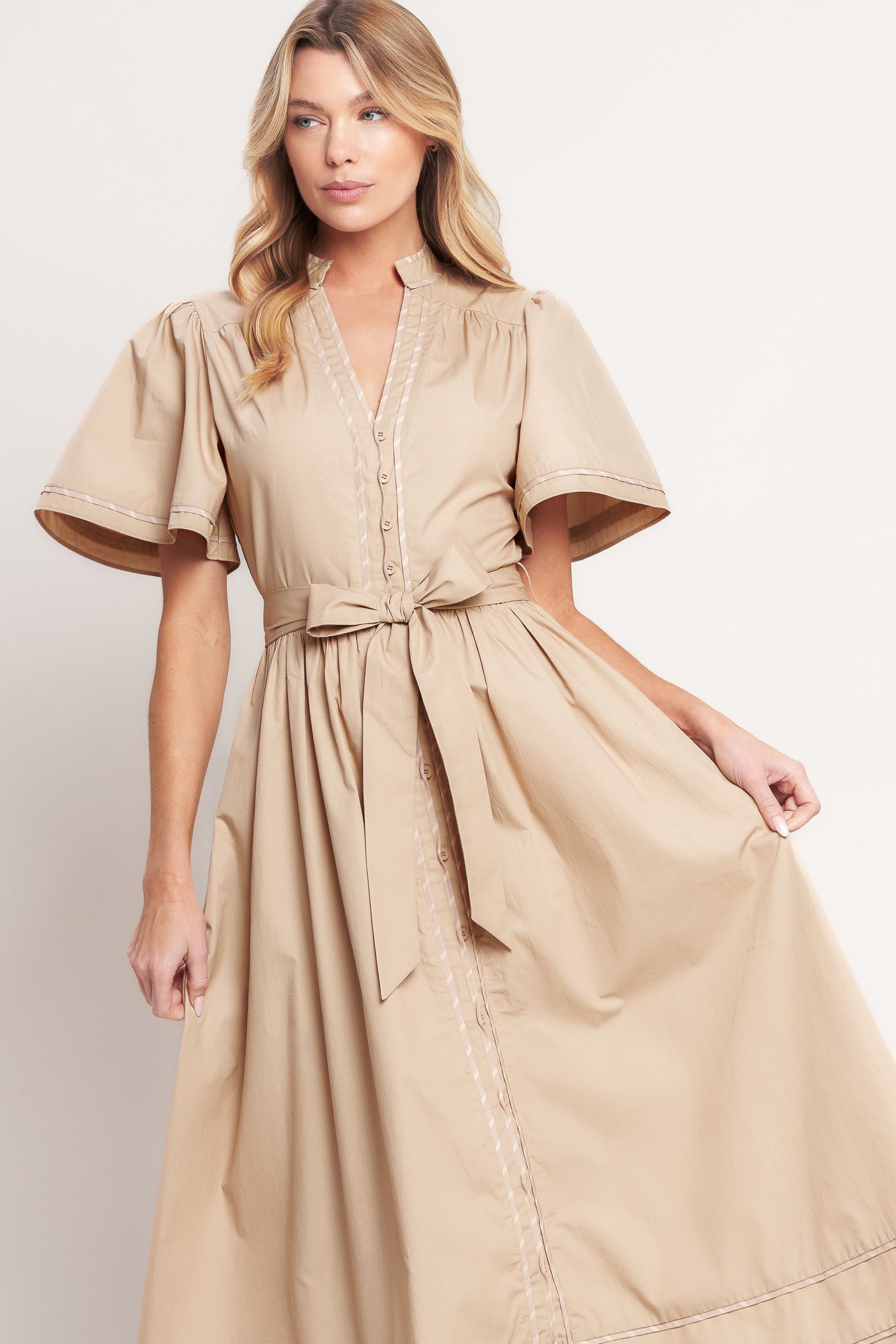 Woman wearing a beige midi dress featuring V neckline, short flutter sleeve, button down, self sash tie, contrasting binding and full skirt | Flying Tomato