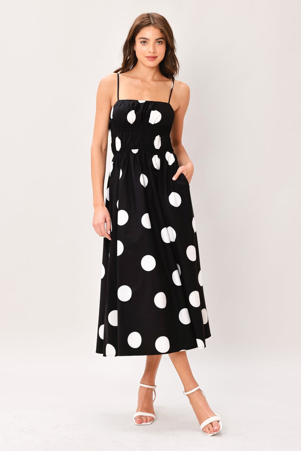 Woman wearing a black midi dress with white polka dots featuring straight neckline, straps, elastic at empire and waist, side pockets and full skirt | Flying Tomato